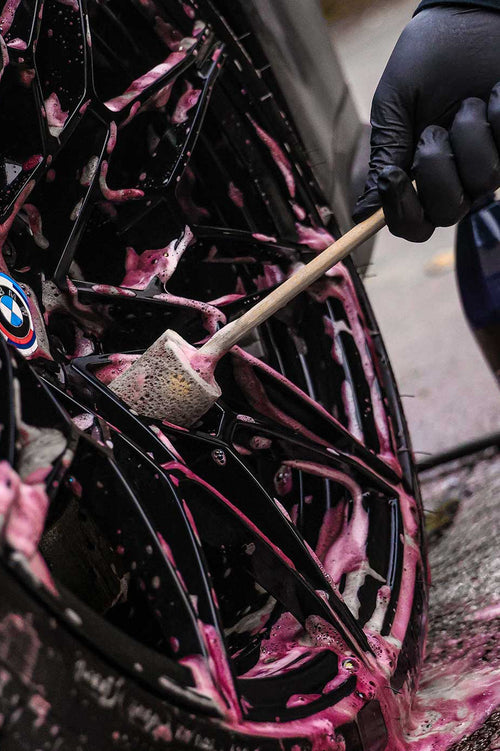 Person cleaning a car tire with pink soap and a brush, wearing black gloves.