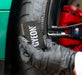Hand wearing a grey glove holding a Gyeon tyre dressing applicator near a car tire and red brake caliper.