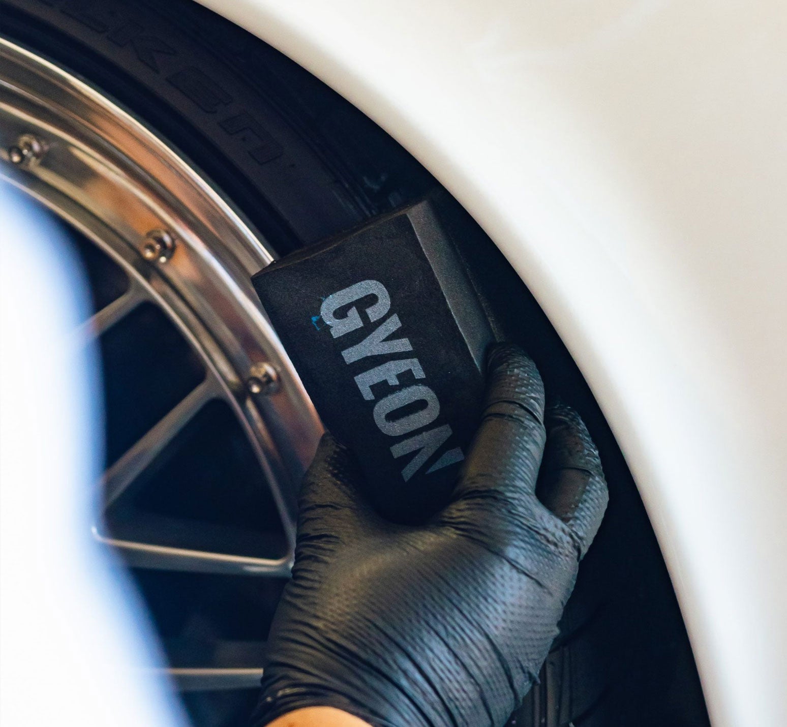 Person wearing a black glove holding a GYEON tyre dressing applicator near a car wheel.