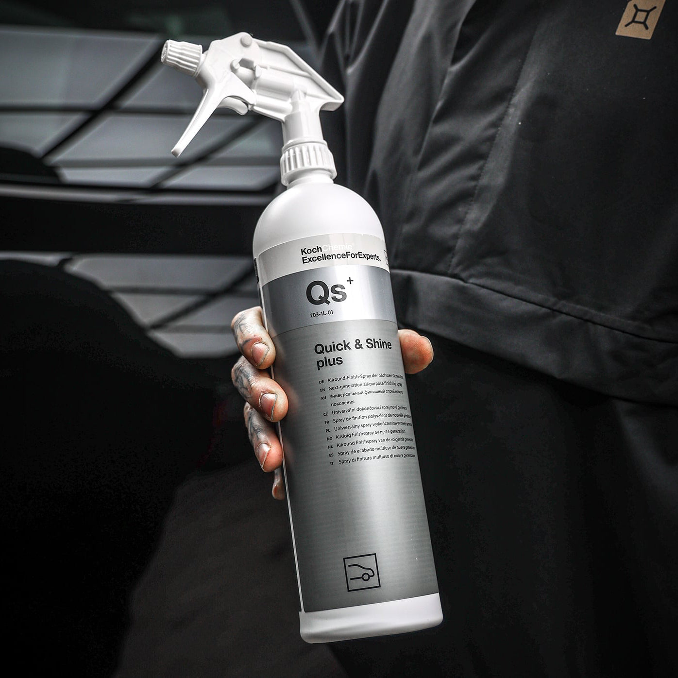 A person in a black uniform holds a spray bottle of Qs+ Quick & Shine Plus car polish. The bottle is white and silver with a trigger spray nozzle, set against a dark, blurred background.