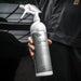 A person in a black uniform holds a spray bottle of Qs+ Quick & Shine Plus car polish. The bottle is white and silver with a trigger spray nozzle, set against a dark, blurred background.