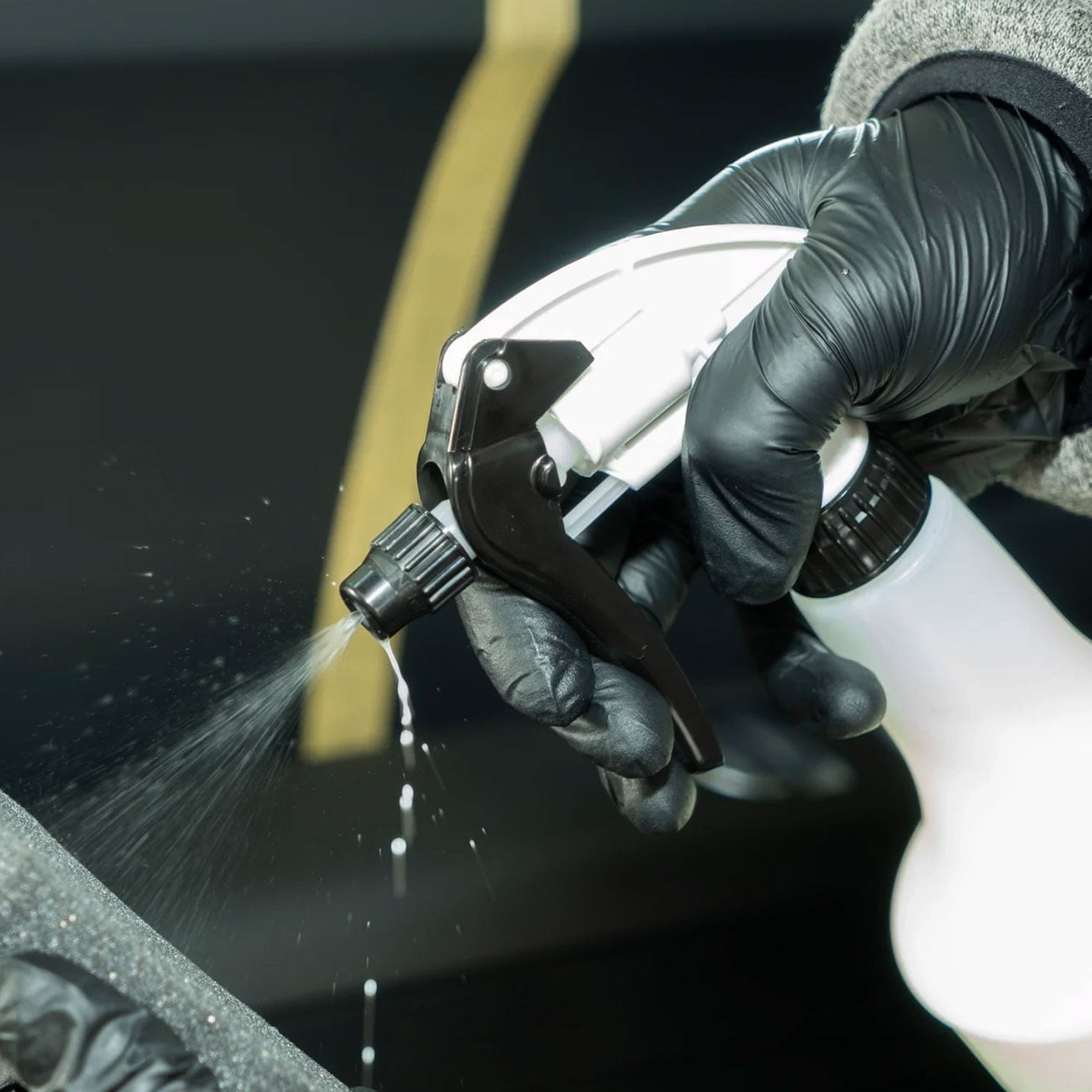 A close-up of a hand wearing a black nitrile glove holding a white spray bottle with a black trigger nozzle, spraying a fine mist. The background is a dark, out-of-focus surface with a yellow stripe.